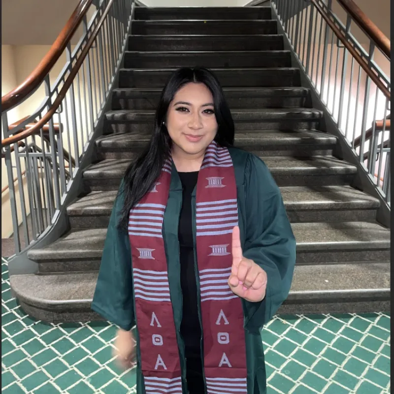 Woman holding her hand up in a number one sign standing by a staircase wearing a green graduation gown and greek stole