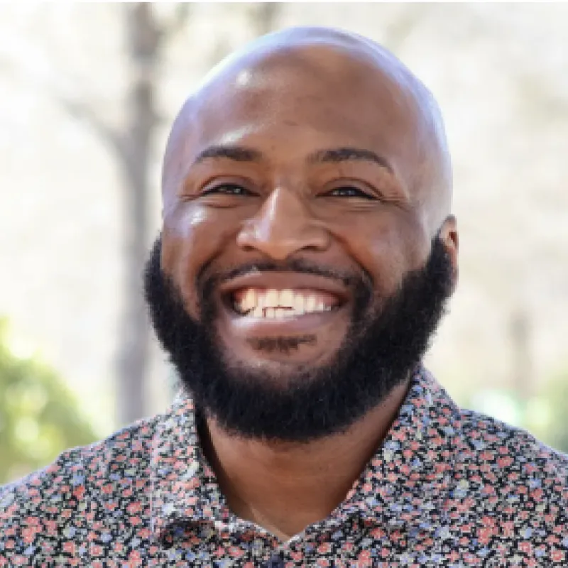 Black, bald man with a full beard smiling outside. He is wearing a floral collared shirt