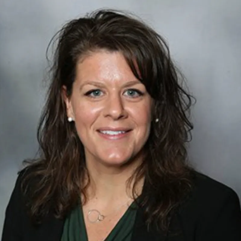 Woman with dark brown hair smiling in front of a gray background. She has blue eyes and pearl earrings with a black blazer and green blouse.