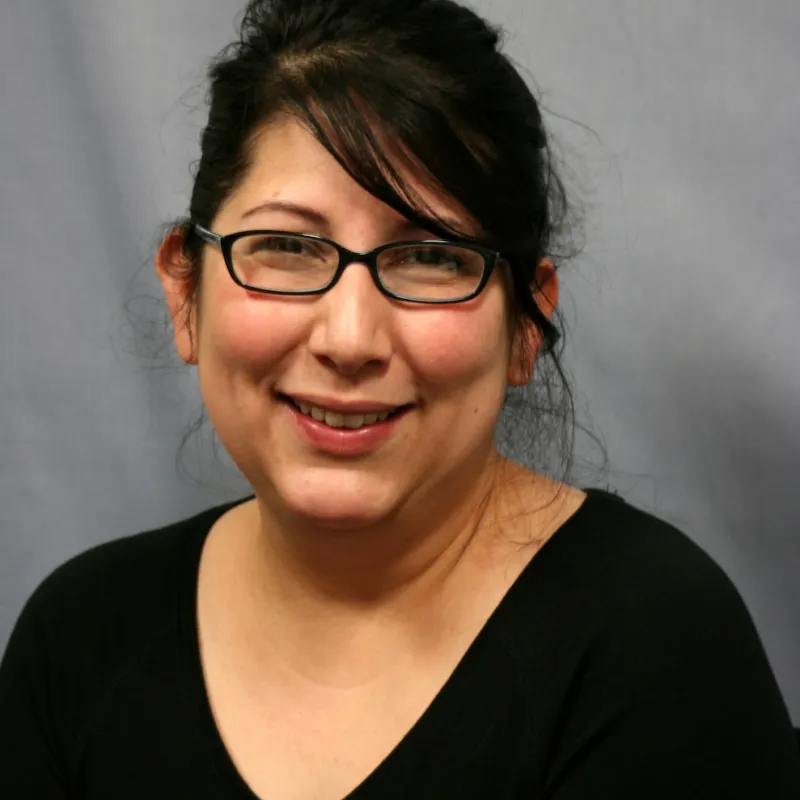 SMiling woman with dark hair and bangs sweeping to the right of her face. She is wearing dark rectangular glasses and a black scoop neck top