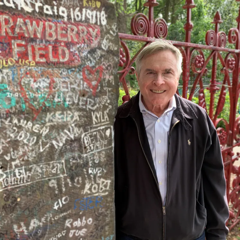 Man with light hair leaning against concrete wall wearing a button up shirt and a black coat