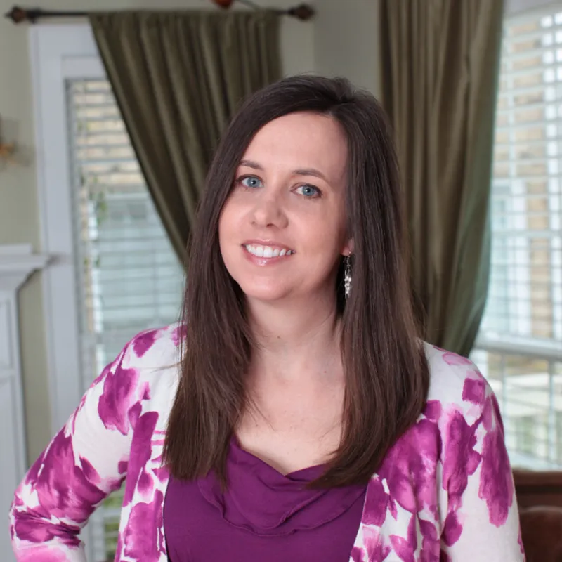 Woman with long brown hair smiling indoors. She is wearing a purple blouse and a pink and white sweater.