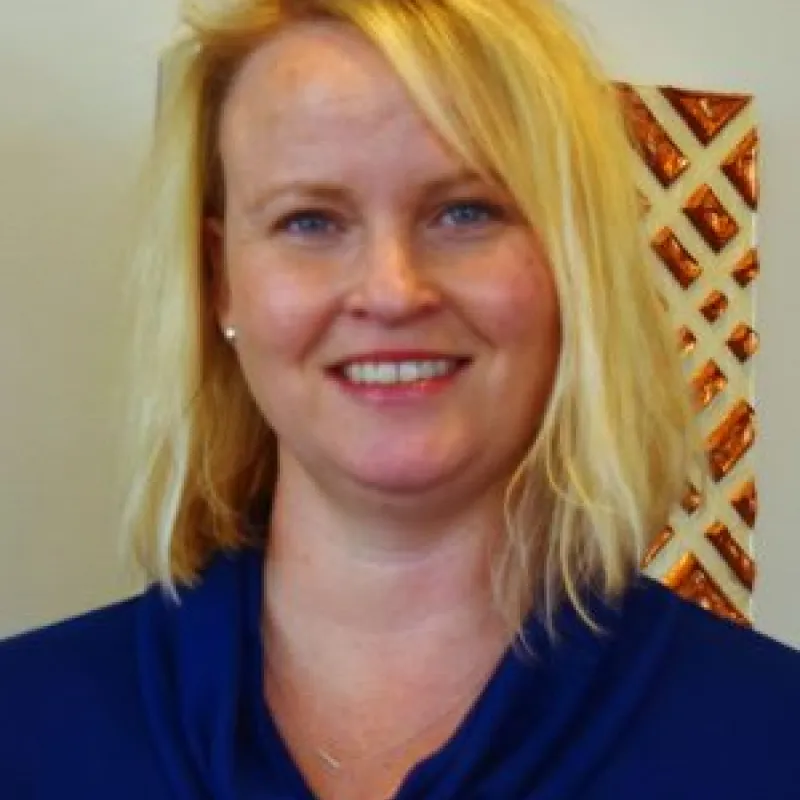 Smiling woman with shoulder-length blonde hair and a dark blue blouse standing in front of a wall