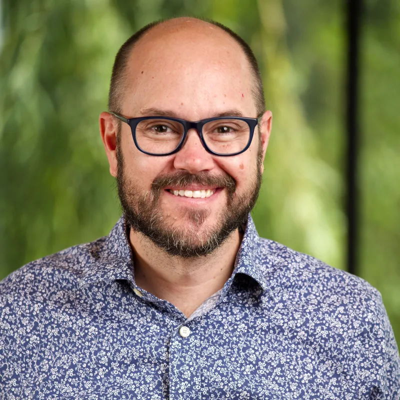 Man smiling in front of a green background. Wears glasses, is brunette with a beard, and is wearing a blue patterned button down shirt.