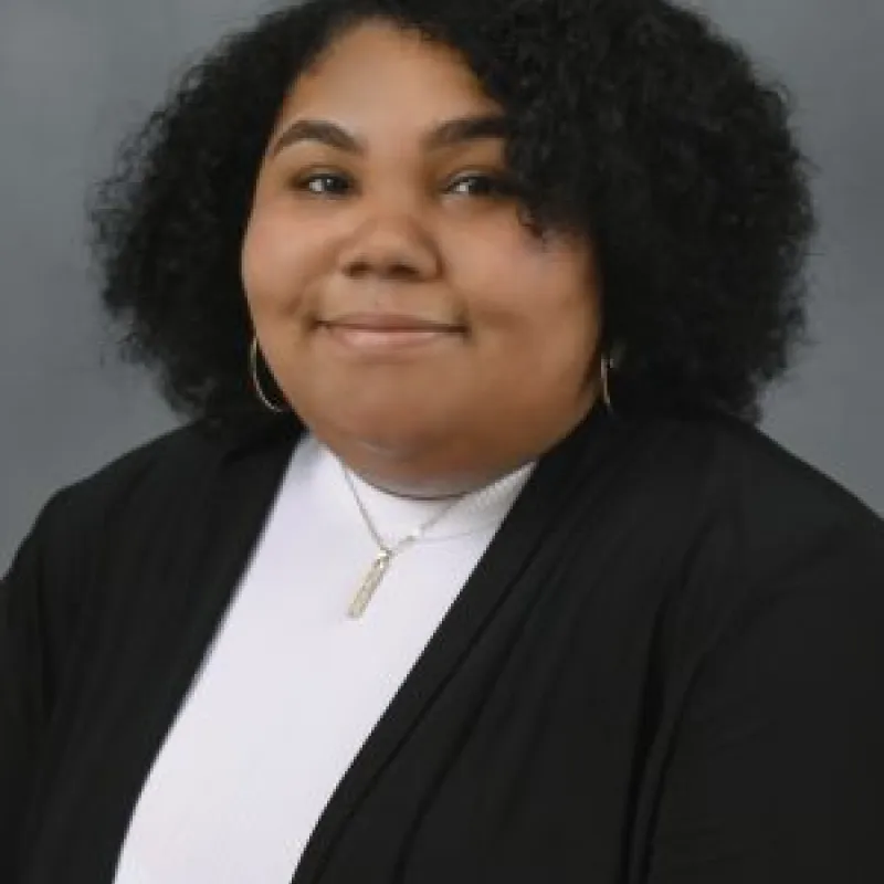 Brown woman with curly, dark hair smiling in front of a gray background. She is wearing gold earrings and a necklace with a white top and a black blazer.