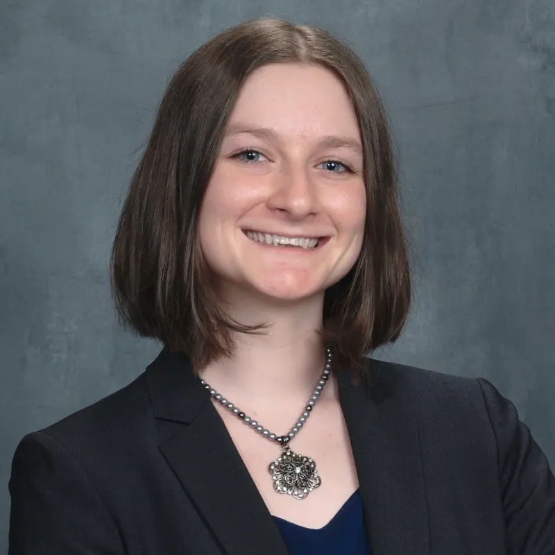 Person with short brown hair wearing a black blazer paired with a flower necklace