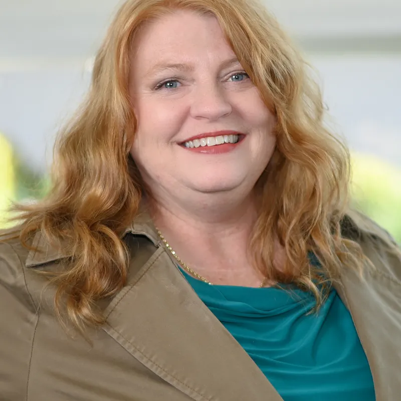 Woman with blond wavy hair smiling wearing a blue top with a buttoned khaki blazer