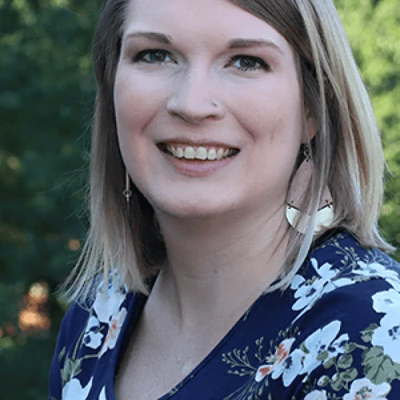 Woman with light skin smiling outside with blonde,, shoulder-length hair. She has a nose piercing, a blue and white floral top and tear drop shaped dangling earrings.