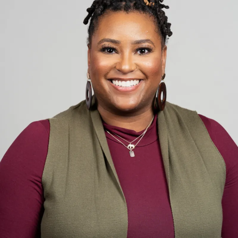 Black, female presenting person with dark to light locs smiling in front of a gray background. Wearing a burgundy long sleeve short with a green vest, dangly, circular earrings, and a necklace. 