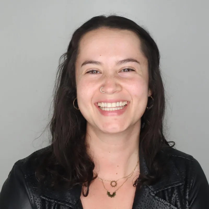Woman with medium length, dark, brown hair smiling in front of a plain, white background. She is wearing a black top with a black leather jacket over it and two dainty necklaces. She has a silver nose ring on the right nostril.