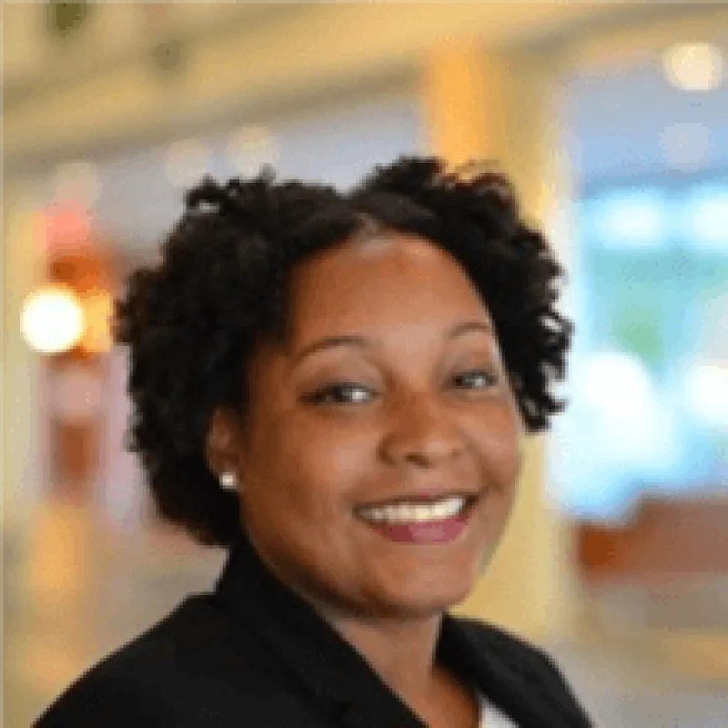 Person with short, black hair parted in the middle and pearl earrings smiling with teeth. They are wearing red lipstick, a black blazer, and a white shirt.