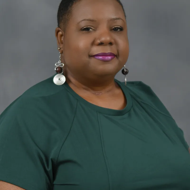 Woman with buzzed hair, dangling earrings, and an emerald green blouse in front of a gray background