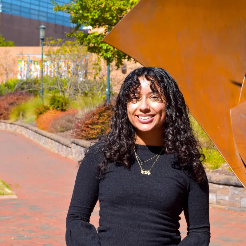 Person standing outside with long, dark curly hair, a gold name necklace, and a black long sleeve shirt. The person has tan skin, a big smile, and dimples. They are outside near a fountain on a sunny day.