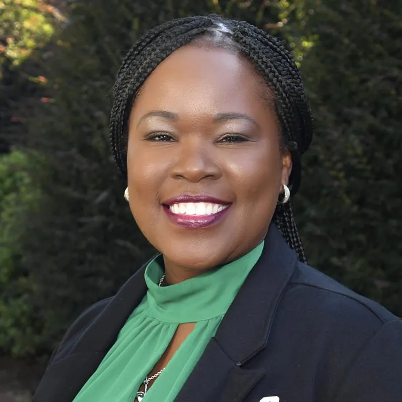 Black woman smiling with a berry lipstick color, silver earrings, and braids pulled back. Wearing a green, mock-neck blouse with a keyhole and a black blazer over it. There are trees in the background with sunshine.