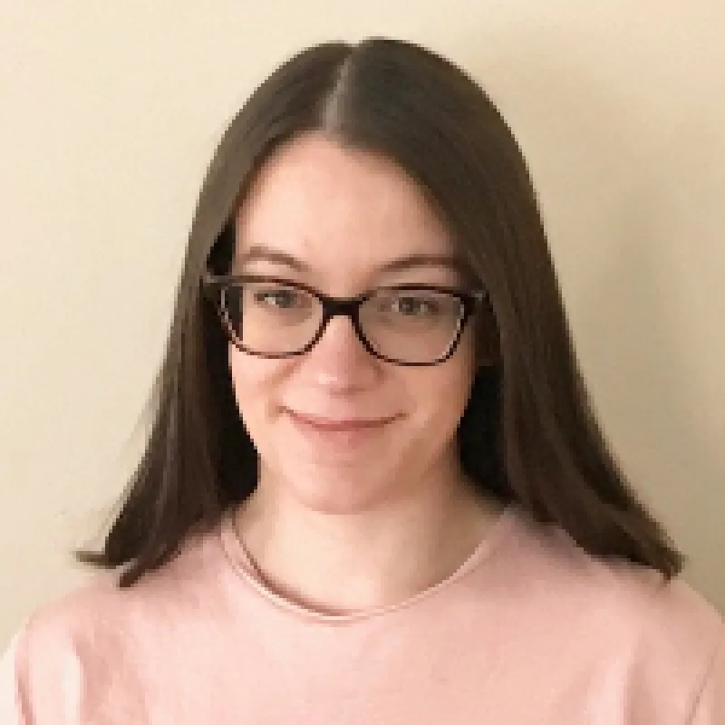 Woman with straight brown hair and glasses smiling in front of a beige background. She is wearing a pink, scoop neck top. 