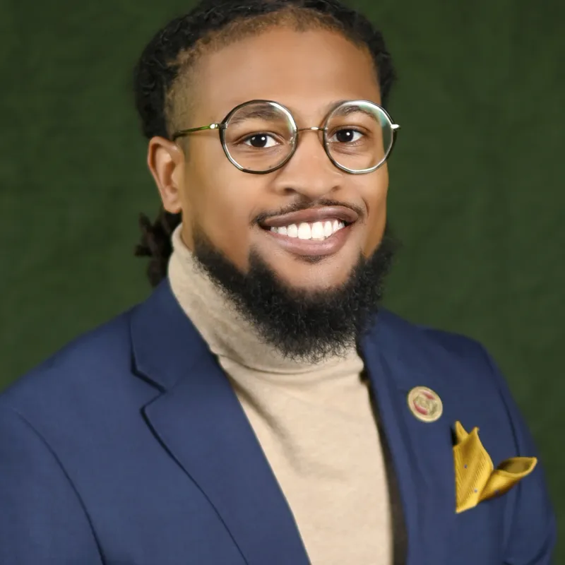 Man wearing round glasses and blue blazer, has black hair and black beard