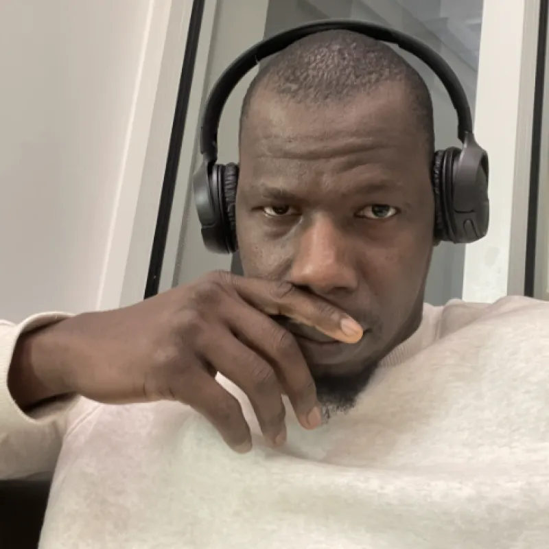 Black man with a buzzcut, black over-ear headphones and a heather gray crewneck. He is sitting in a building in front of a window and has his hand covering his mouth