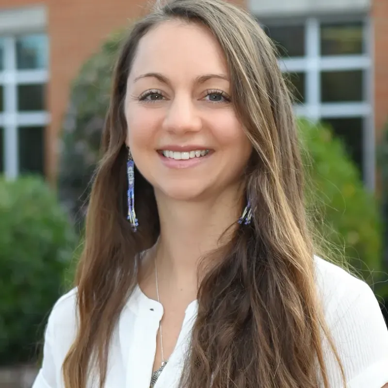 woman with long brown hair, dangling earrings, and a white blouse smiling in front of a brick building