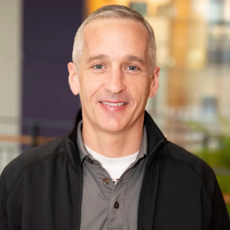 Man with short gray/white hair smiling in a building. He has a gray shirt on with a black light jacket over it