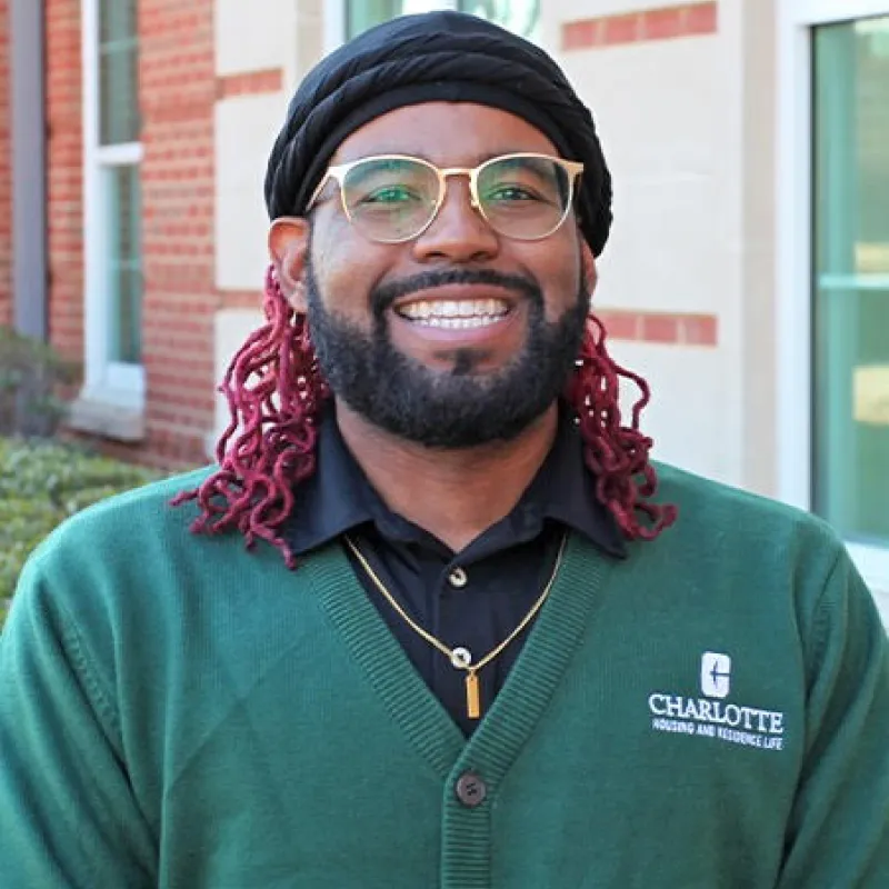 Person standing outside smiling with a Charlotte green cardigan over a black polo. They have a black turban over red curly locs, and have a full beard and mustache. They are also wearing a gold necklace and gold glasses.