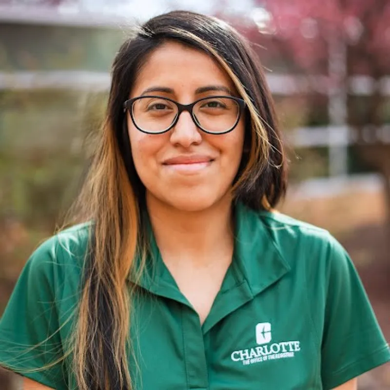 Woman with black glasses, closed mouth smile, and long brown hair standing outside wearing a green UNC Charlotte polo