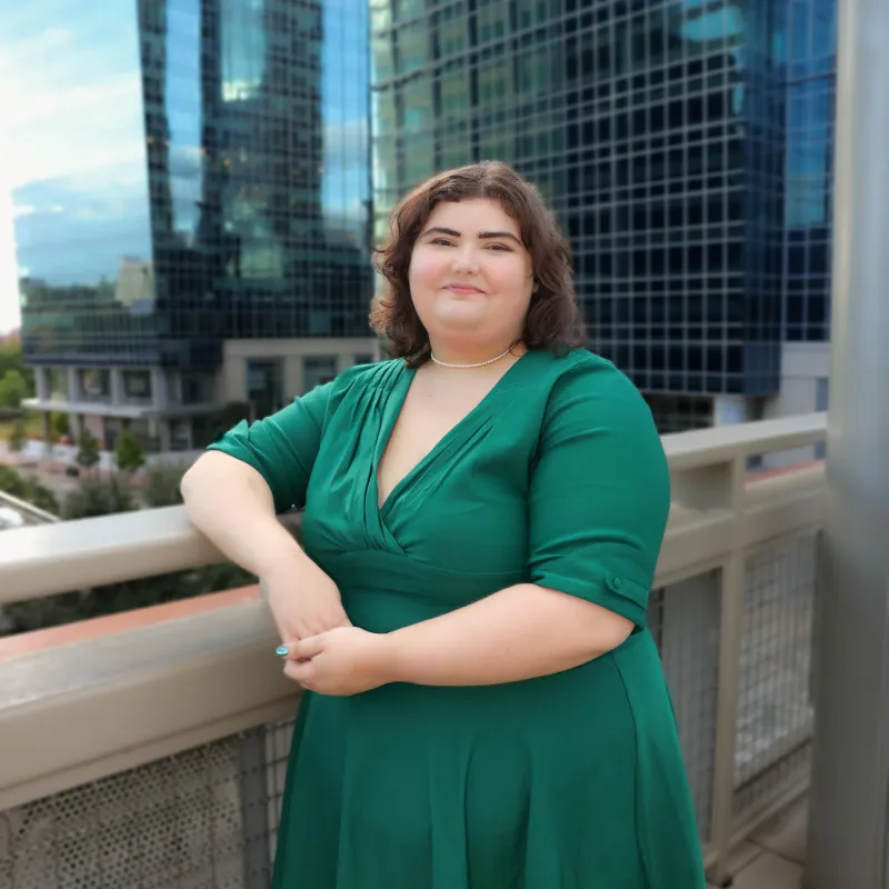 Person with short, brown hair smiling in front of skyscrapers. They are wearing a green dress, makeup, and is leaning on a railing.