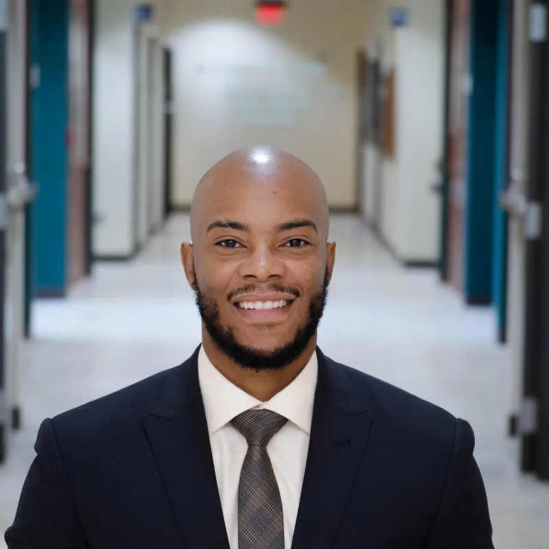 Bald Black man smiling wearing a dark suit, white button down, and a grey tie. He has a beard and is in a hallway of a building